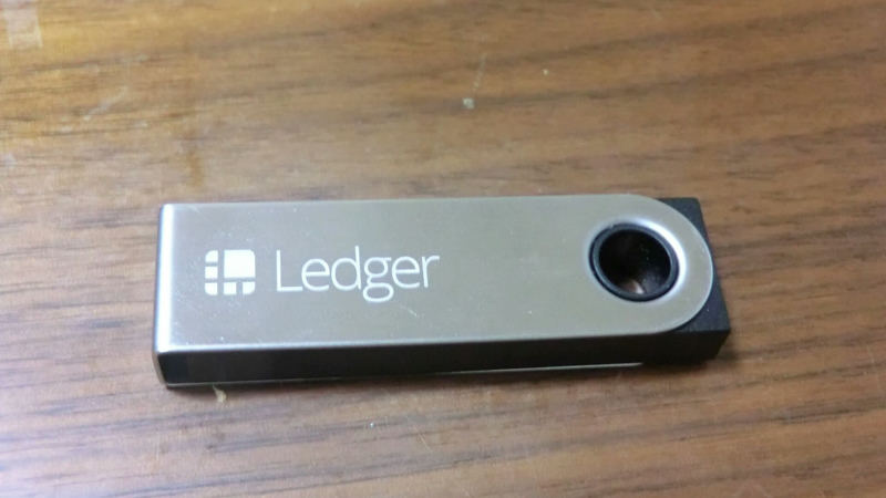 A small Ledger Nano S hardware wallet, shaped like a USB stick with a metal case, resting on a wooden desk.