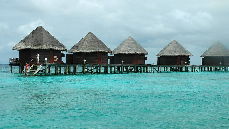 A row of thatched water bungalows on stilts above a shallow turquoise lagoon at Thulhagiri Island Resort in the Maldives.