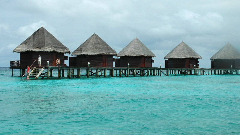 A row of thatched water bungalows on stilts above a shallow turquoise lagoon at Thulhagiri Island Resort in the Maldives.