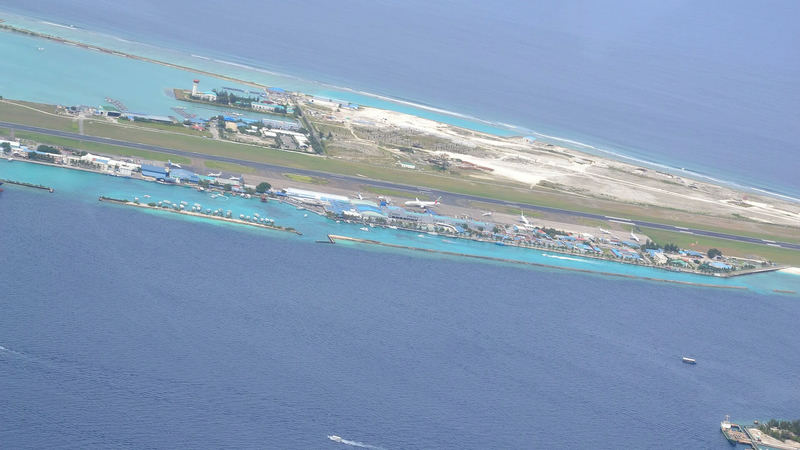 An aerial view of Velana International Airport: a long runway and terminal buildings on a narrow island surrounded by turquoise sea.
