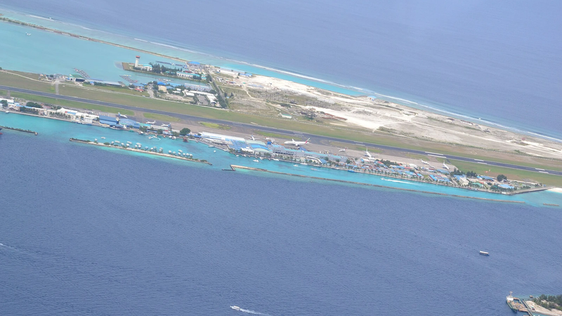 An aerial view of Velana International Airport: a long runway and terminal buildings on a narrow island surrounded by turquoise sea.