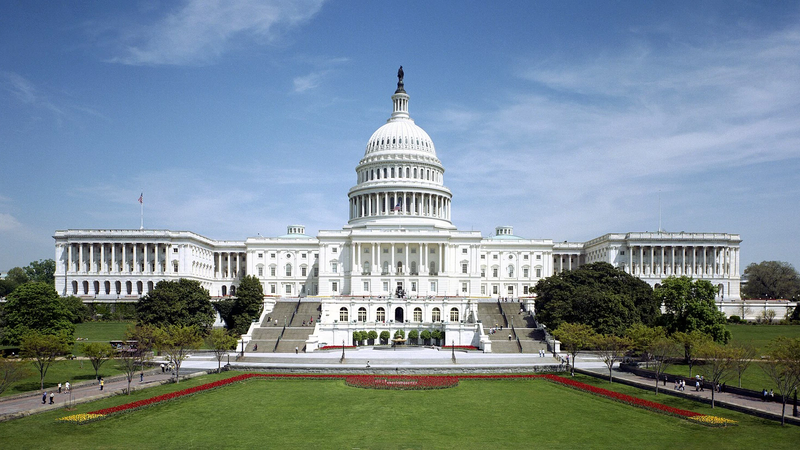 The west front of the United States Capitol building in Washington, DC, seen across green lawns and flower beds under a blue sky.