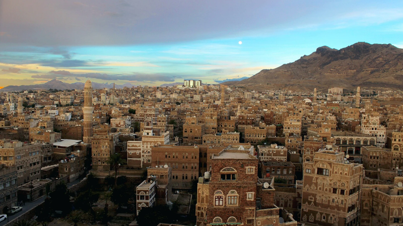 A view over the Old City of Sanaa in Yemen at dusk, with its tall decorated tower houses and mountains in the background.
