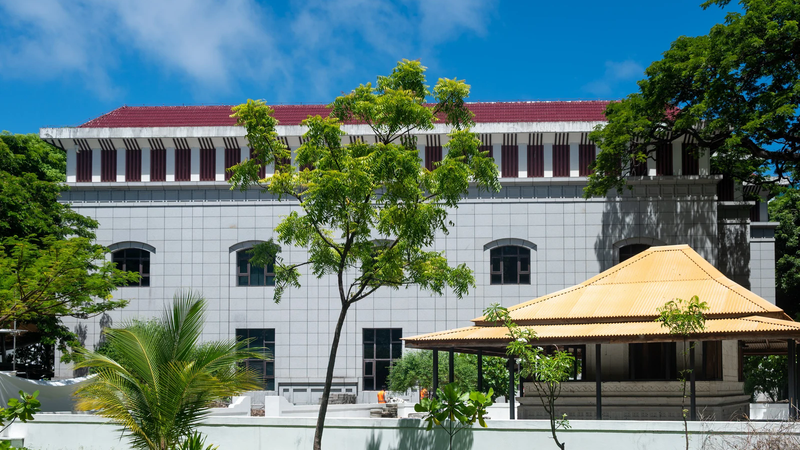 A whitewashed building with a red roof beside a yellow-roofed pavilion and a lawn in Malé