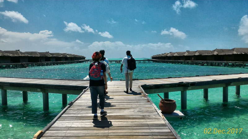 Visitors walking along a wooden jetty towards rows of thatched water villas over a turquoise lagoon