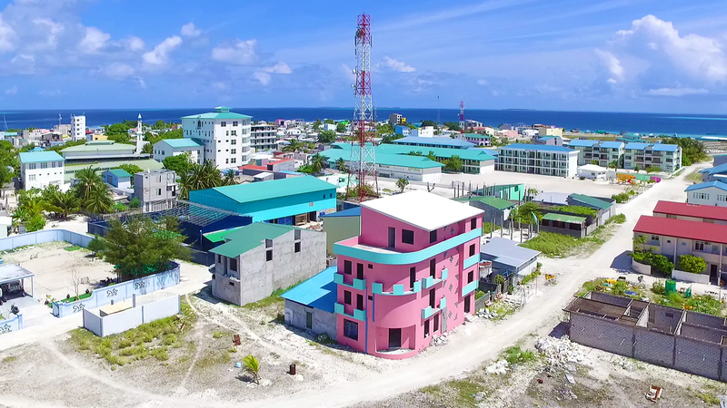 Aerial view of colourful houses, low-rise buildings and a telecommunications mast on Naifaru island, Lhaviyani Atoll, with the ocean behind