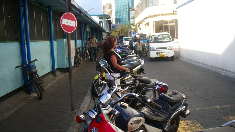 A long row of motorcycles parked along the edge of a narrow street in Malé, with buildings and a no-entry sign nearby.