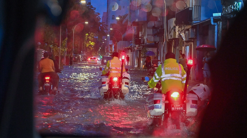 Police on motorcycles ride through floodwater on a street in Malé at night during heavy rain, with people sheltering under umbrellas.