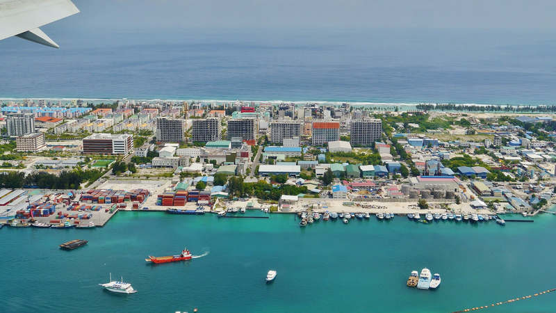 An aerial view of Hulhumalé showing apartment blocks, the harbour with boats and containers, and open undeveloped land near the shore.