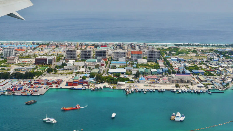 An aerial view of Hulhumalé showing apartment blocks, the harbour with boats and containers, and open undeveloped land near the shore.