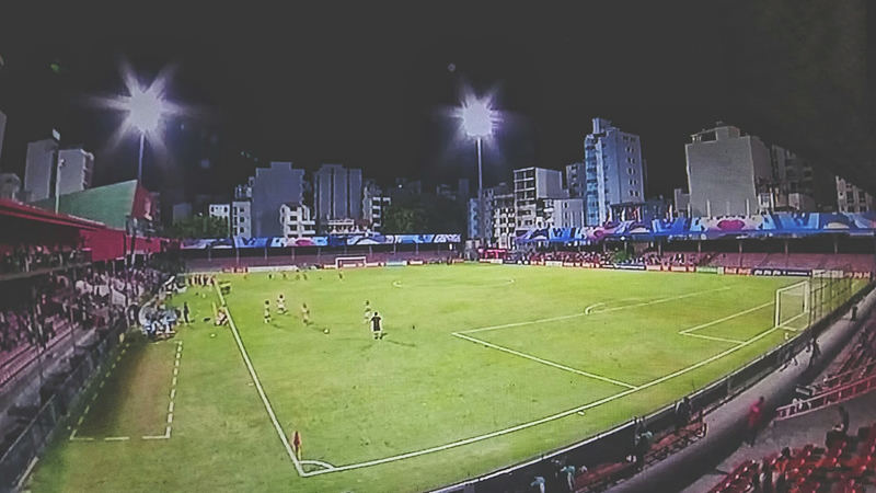 Floodlit football pitch at the National Stadium in Malé with city buildings behind the stands