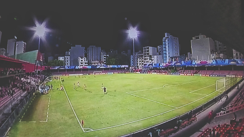 Floodlit football pitch at the National Stadium in Malé with city buildings behind the stands