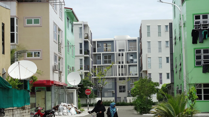 Two women walking across a quiet street in Hulhumalé lined with apartment blocks, a motorcycle parked at the kerb