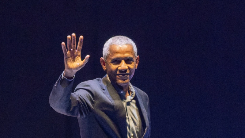 Barack Obama in a dark suit smiles and raises his hand to wave to the audience on a dimly lit stage