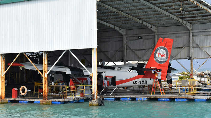Two Trans Maldivian Airways seaplanes under maintenance at a floating dock beside turquoise water, with engineers working on the red and white tail of one aircraft