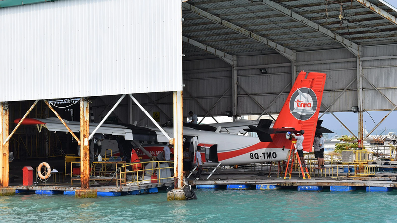 Two Trans Maldivian Airways seaplanes under maintenance at a floating dock beside turquoise water, with engineers working on the red and white tail of one aircraft
