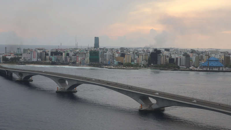 An aerial view of the Sinamalé Bridge curving across the sea towards the dense buildings of Malé at dusk