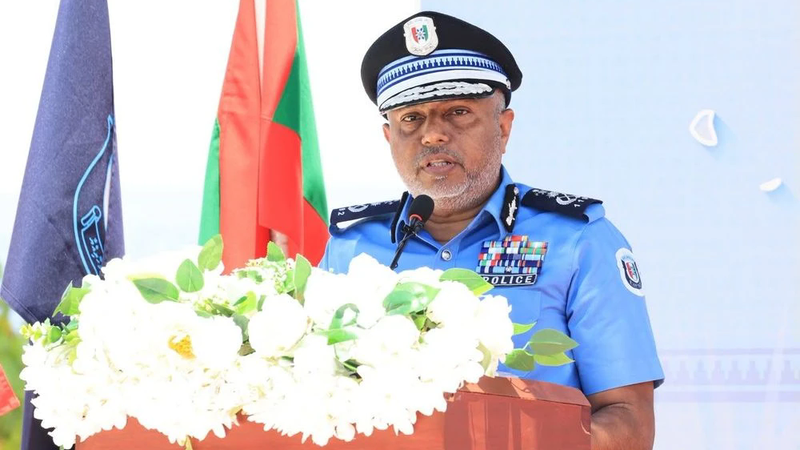 Commissioner of Police Ahmed Mohamed in a blue uniform and peaked cap speaking at a podium decorated with white flowers, with the Maldivian flag behind him