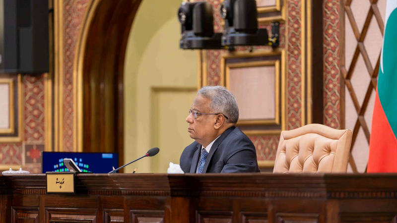 The presiding officer seated at the chair's desk in the chamber of the People's Majlis, with the Maldivian flag behind.