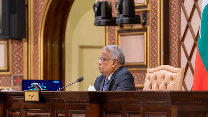 The presiding officer seated at the chair's desk in the chamber of the People's Majlis, with the Maldivian flag behind.