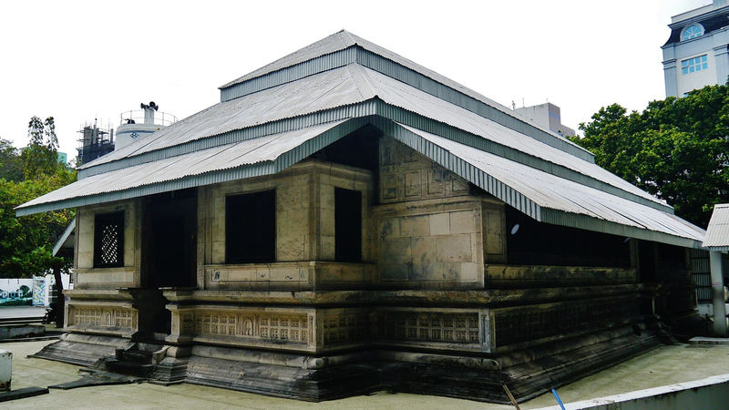A low coral-stone mosque with carved walls and a tiered grey metal roof, with trees and modern buildings behind it in Malé.