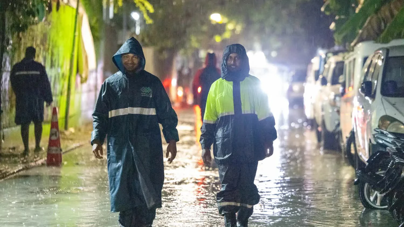 Two men in hooded raincoats walk through a flooded street at night in heavy rain, with parked cars and motorbikes on one side.