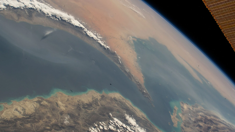 View from space of a narrow sea passage between desert coastlines, with the curve of the Earth and a solar panel at the top right.