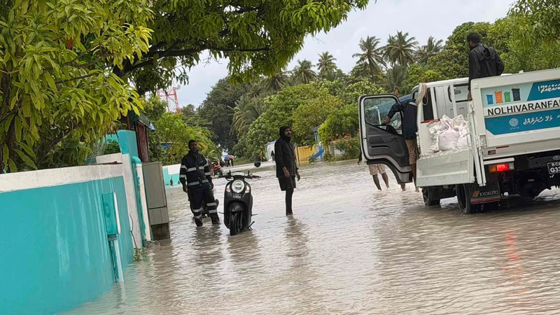 Floodwater covers a residential street lined with trees, with a police officer, residents and a council truck loaded with sandbags