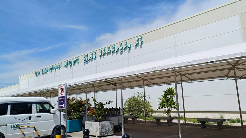 Front of the Gan International Airport terminal building with its name sign in English and Dhivehi, a covered walkway and a parked van