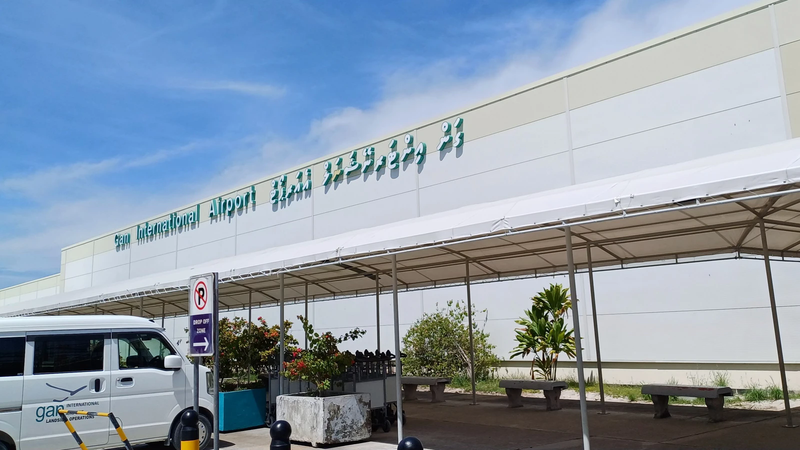 Front of the Gan International Airport terminal building with its name sign in English and Dhivehi, a covered walkway and a parked van