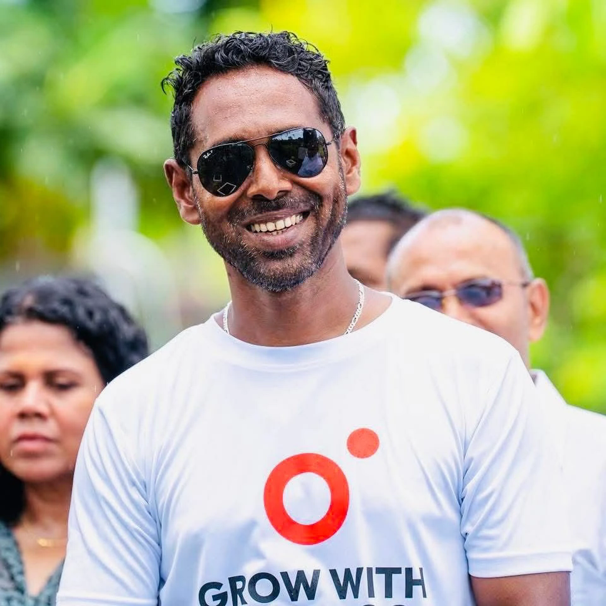 Abdullah Ramzy smiling in sunglasses and a white T-shirt, with other people and green trees behind him