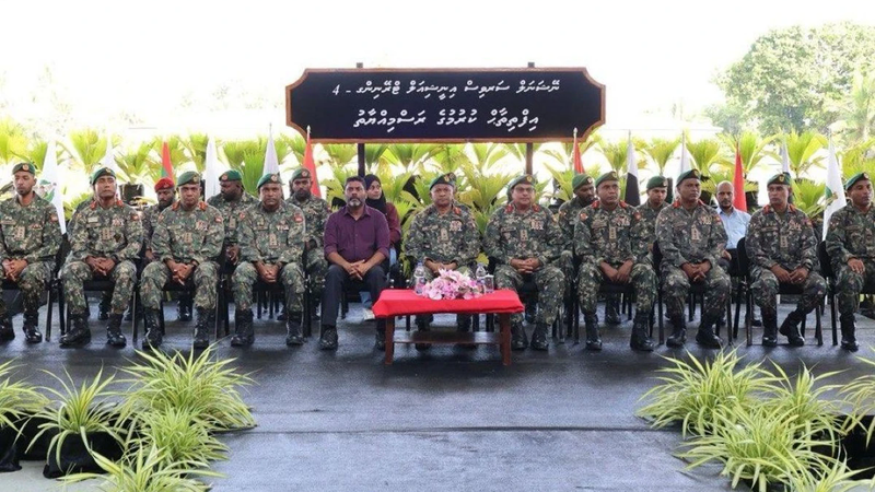 Rows of MNDF officers in camouflage uniforms and green berets seated on a stage beneath a Dhivehi sign for the national service training ceremony