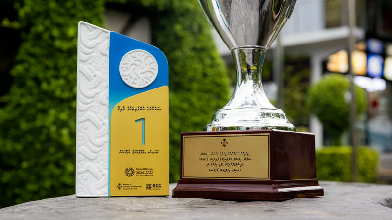 A silver trophy on a wooden base beside a white, blue and yellow first-place award plaque from Maldives Expo 2026, set outdoors against green foliage