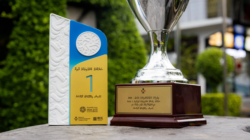 A silver trophy on a wooden base beside a white, blue and yellow first-place award plaque from Maldives Expo 2026, set outdoors against green foliage