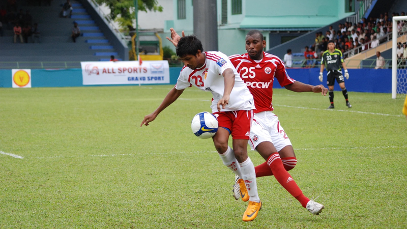 Two footballers, one in a white kit and one in red, compete for the ball on a grass pitch in front of a stand