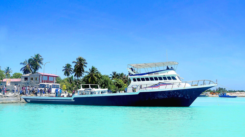 A large dark-hulled fishing vessel moored beside a harbour quay with palm trees and people on shore, in turquoise water