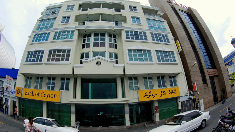 Street view of a white bank building with yellow Bank of Ceylon signs next to the Bank of Maldives tower in Malé, with taxis and motorcycles on the road