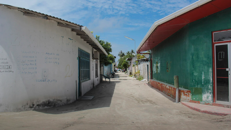 A quiet sandy street between houses on a Maldivian island, with a parked motorcycle in the distance