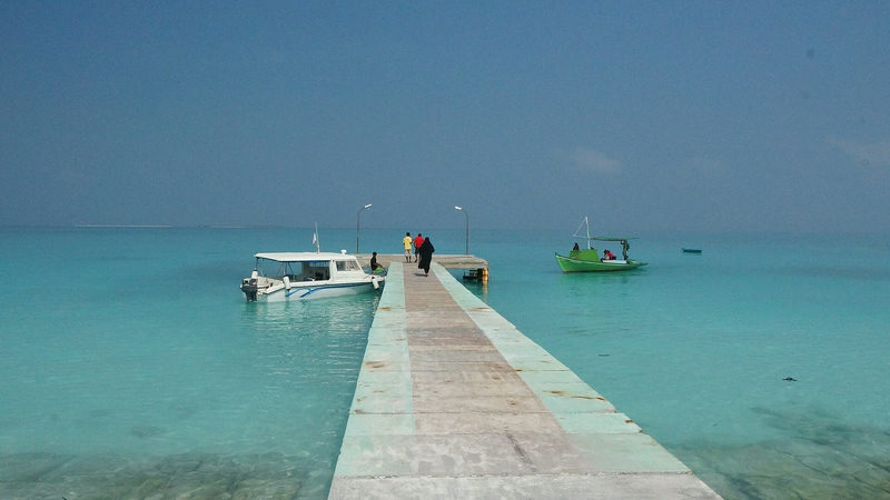 A long concrete jetty leading into turquoise water, with a speedboat and a small green boat moored alongside and people walking towards the end