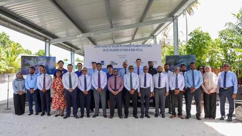 A group of officials in shirts and ties stand in a row under a metal canopy in front of a banner about launching solar PV hybrid power systems in Raa and Baa atolls