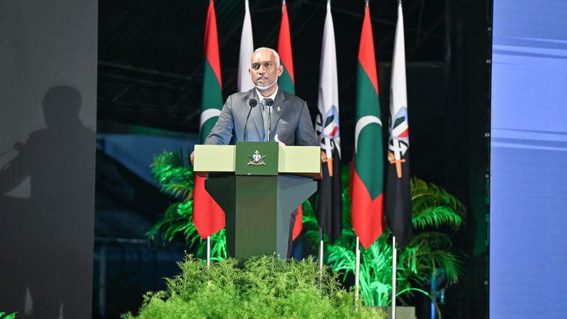 President Mohamed Muizzu speaking at a green podium in front of Maldivian and MNDF flags at the MNDF anniversary ceremony