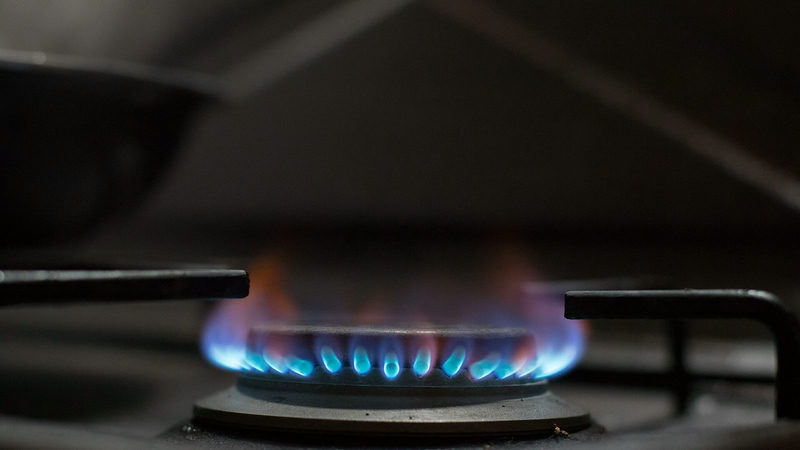 Close-up of blue flames from a gas stove burner in a dark kitchen, with a pan blurred in the background