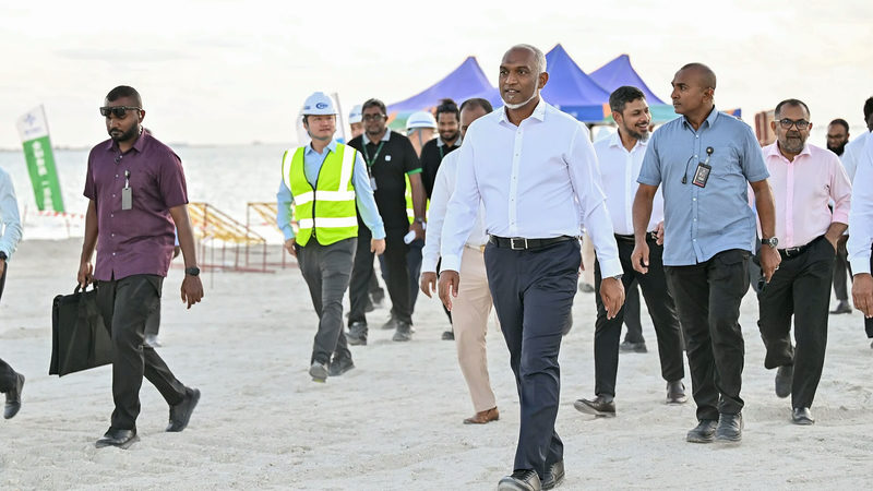 President Mohamed Muizzu walks on white reclaimed sand at the Ras Malé site with officials and a worker in a hard hat