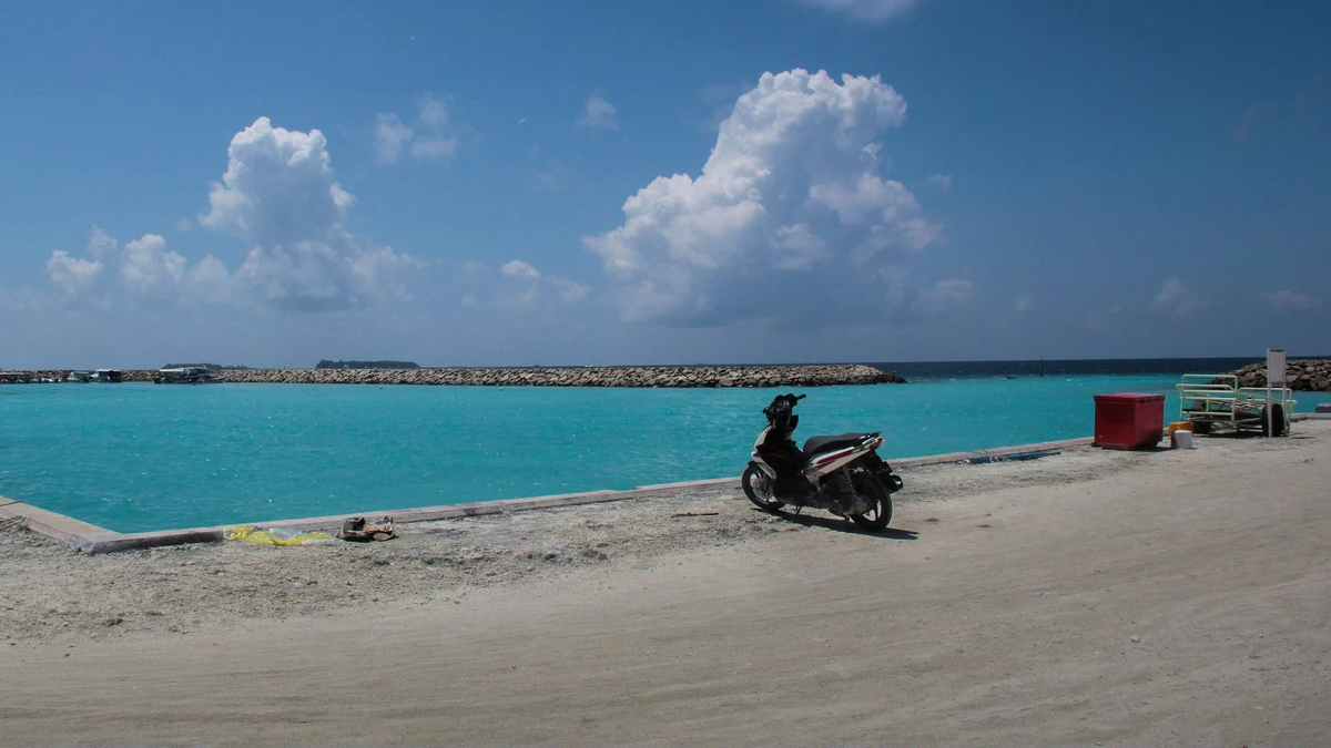 Port area of Maafushi, Maldives, with a parked scooter on the quay, turquoise water and a rock breakwater under a blue sky