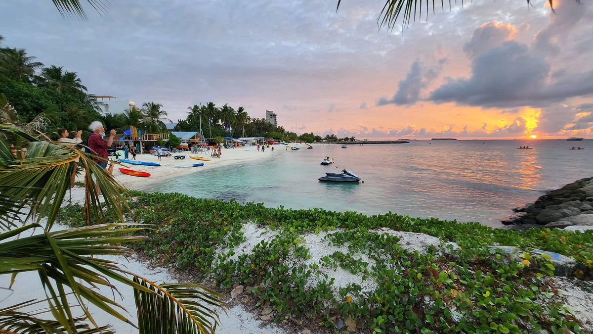 Sunset over the water sports beach in Maafushi, Maldives, with kayaks on the sand, jet skis in the lagoon and visitors taking photos