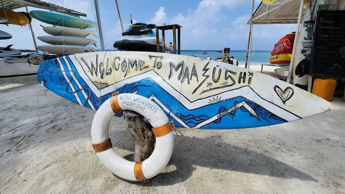 Painted surfboard reading Welcome to Maafushi with a lifebuoy, jet ski and paddleboards on a beach in Maafushi, Maldives