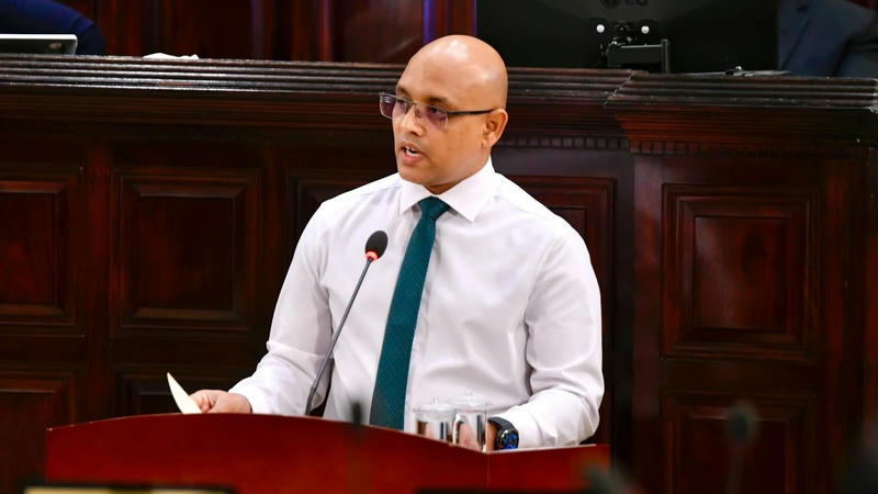 Finance Minister Hassan Zareer, in a white shirt and green tie, speaks at a wooden podium in the Majlis chamber