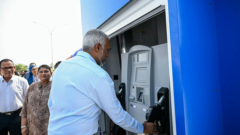 A man in a light blue shirt holds a fuel nozzle at a blue STO fuel dispenser as onlookers watch
