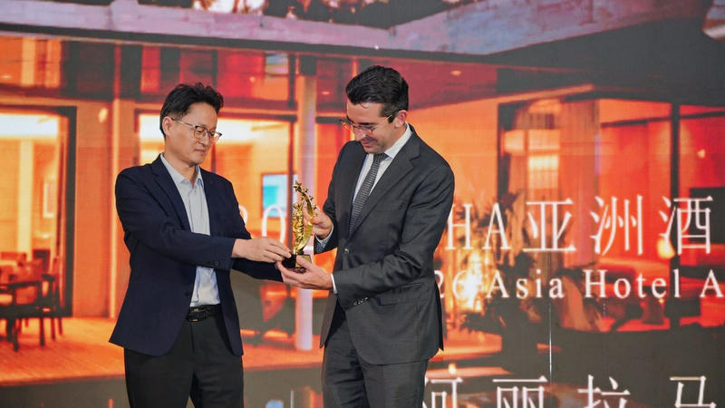 Two men in suits hold a gold trophy on stage in front of a screen showing the Asia Hotel Awards and a resort villa